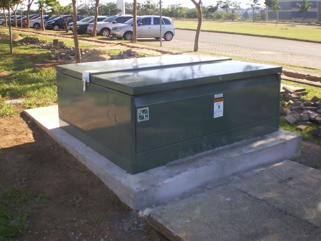 The fully installed Vista switchgear unit, complete with its dark green S&C-branded enclosure, sits on its concrete pad in the foreground. Evidence of the installation work is present, including a now-empty wood pallet in the foreground, freshly dug dirt around the concrete pad, and scattered materials on the grass. In the background are a row of trees, a row of parked vehicles, and a road.